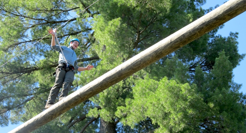 A person wearing safety gear and secured by ropes balances on an obstacle on a high ropes course. 